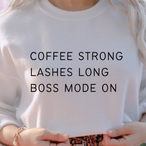 Op de afbeelding: Wit sweatshirt met de tekst "COFFEE STRONG LASHES LONG BOSS MODE ON" gedrukt op de voorkant.