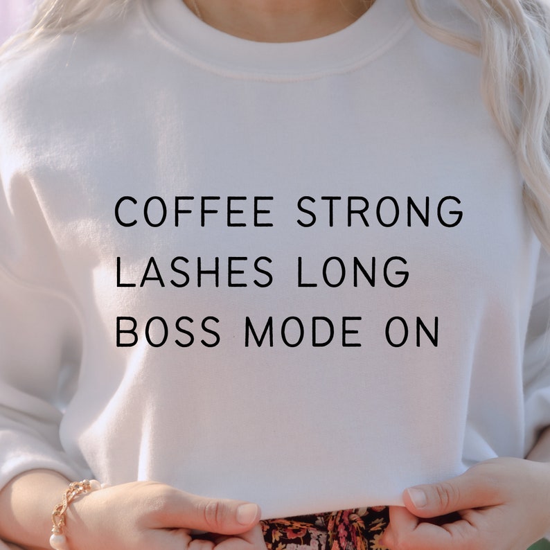 Op de afbeelding: Wit sweatshirt met de tekst "COFFEE STRONG LASHES LONG BOSS MODE ON" gedrukt op de voorkant.