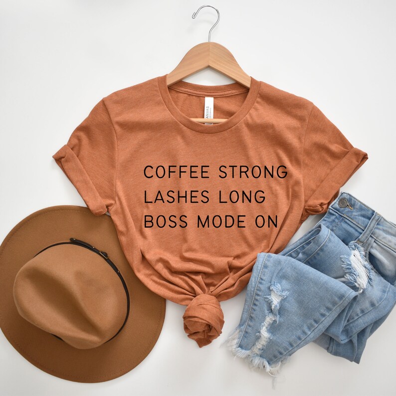 Op de afbeelding: Een roestkleurig T-shirt met de tekst "COFFEE STRONG LASHES LONG BOSS MODE ON" gedrukt op de voorkant.