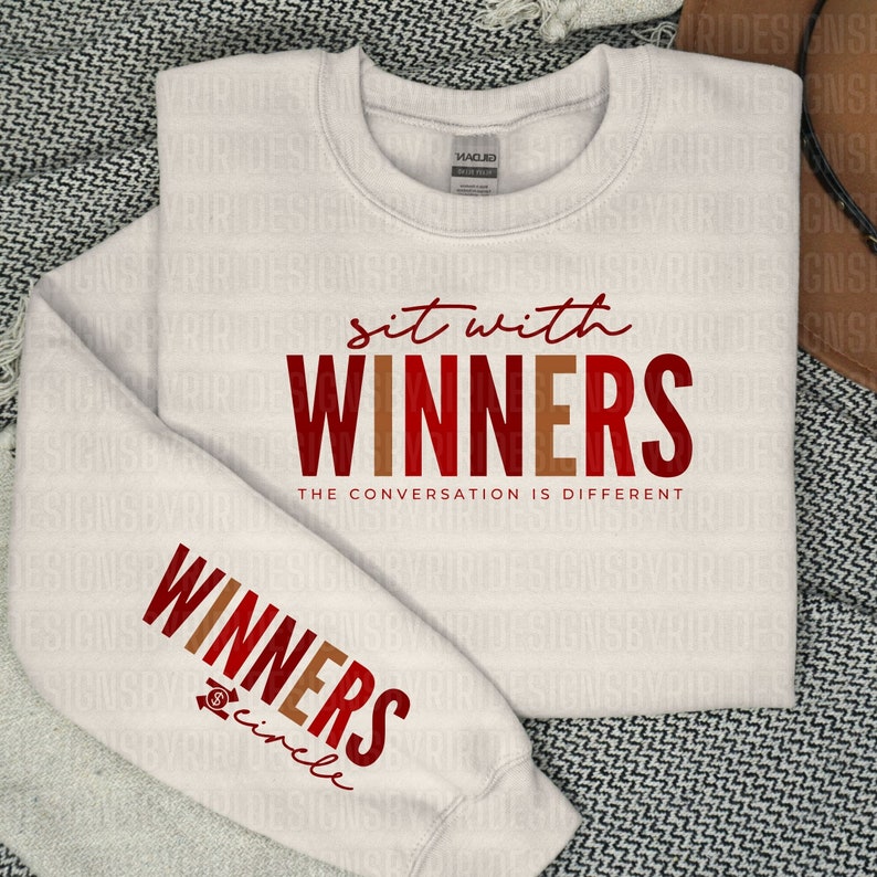 May include: A white sweatshirt with the text "sit with WINNERS" in red and brown lettering. The text "THE CONVERSATION IS DIFFERENT" is below the main text. The left sleeve has the text "WINNERS circle" in red and brown lettering.