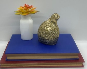 May include: A golden-colored bird figurine with detailed feather textures sits on a blue book. A white vase with a yellow and orange succulent is to the left. A red book is below the blue book.