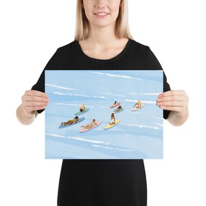 Puede incluir: Una ilustraci&oacute;n de acuarela de un grupo de personas en tablas de surf en el oc&eacute;ano. El agua es azul y el cielo es blanco. Las personas llevan todas trajes de ba&ntilde;o y est&aacute;n remando en sus tablas.