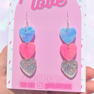 Könnte beinhalten: Ein Paar Hängeohrringe mit drei Herzanhängern. Das obere Herz ist blau-weiß marmoriert, das mittlere Herz ist rosa-weiß marmoriert und das untere Herz ist klar mit schimmerndem Glitzer. Die Ohrringe sind silberfarben und haben einen Hakenverschluss. Die Ohrringe werden auf einer rosa Karte mit dem Text "folksy love" und "www.folkslove.com" und "TAG ME! @folkslove" präsentiert.