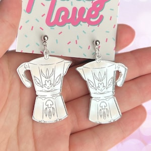 May include: Silver mirrored earrings shaped like a coffee pot with floral designs. The earrings are hanging from silver hooks.