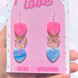 May include: A pair of dangle earrings featuring three heart-shaped charms. The top charm is pink with a swirl pattern, the middle charm is clear with gold glitter, and the bottom charm is blue with a swirl pattern. The earrings are silver and have a hook fastening. The earrings are displayed on a pink and white card with the text "folksy love" and "www.folksylovec.com".
