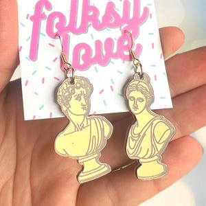 May include: A pair of gold earrings featuring yellow bust sculptures of a man and a woman. The earrings are on a white card with the text "folksy love" in pink.