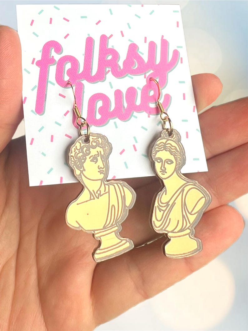May include: A pair of gold earrings featuring yellow bust sculptures of a man and a woman. The earrings are on a white card with the text "folksy love" in pink.