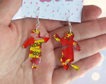 May include: A pair of clear acrylic earrings shaped like stick figures. The figures are covered in red paint splatters and yellow crime scene tape. The tape reads "Police Line Do Not Cross".
