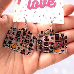 Puede incluir: Un par de pendientes de acrílico transparente con forma de cámara. Los pendientes están decorados con un patrón colorido de cámaras, rollos de película y otros iconos relacionados con la fotografía.