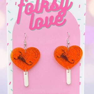 May include: Orange heart-shaped earrings with a black scorpion design and glitter. The earrings are on a white background with the text "folksy love" and "www.folkslove.com" and "TAG ME! @folkslove".
