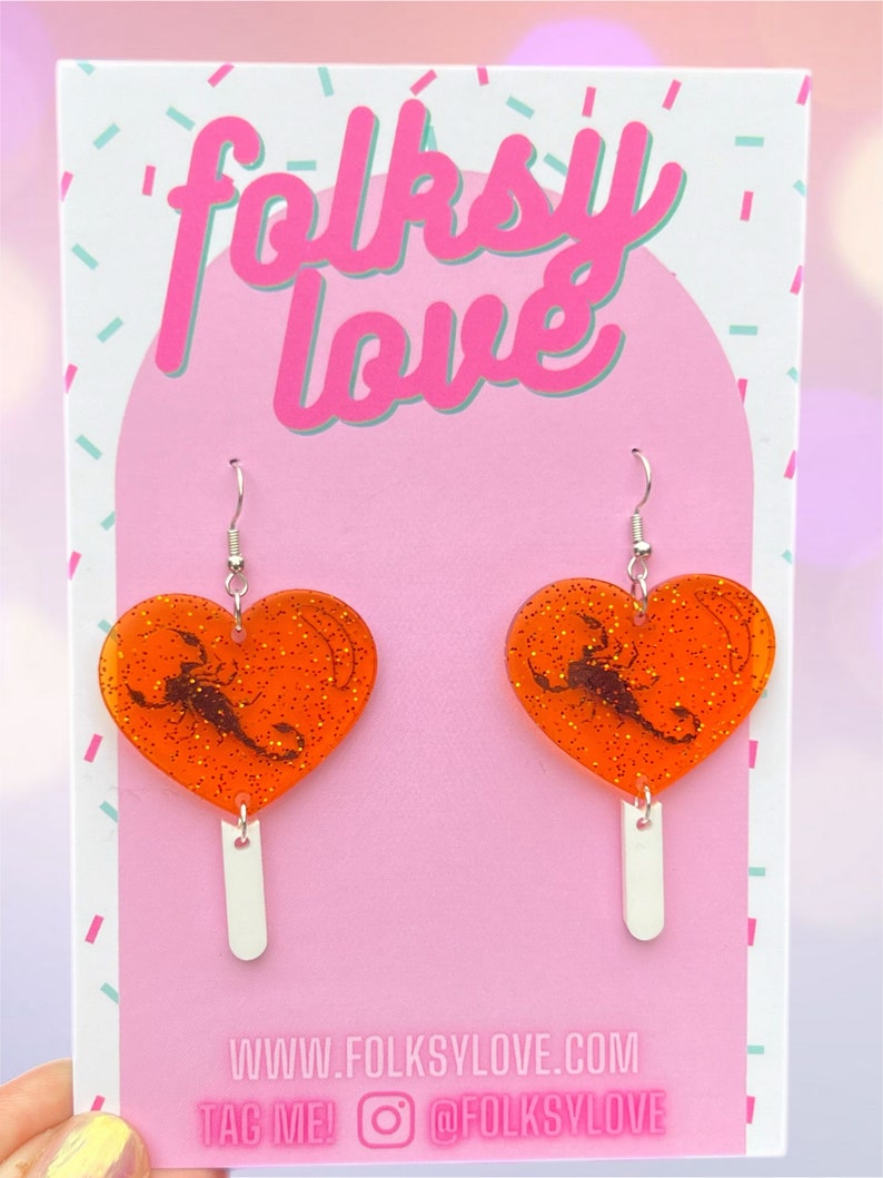 May include: Orange heart-shaped earrings with a black scorpion design and glitter. The earrings are on a white background with the text "folksy love" and "www.folkslove.com" and "TAG ME! @folkslove".