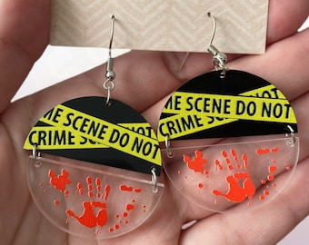 May include: A pair of black and yellow crime scene tape earrings with a red bloody handprint on a clear acrylic background. The earrings are hanging from silver hooks.