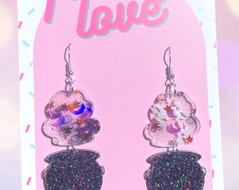 May include: A pair of dangle earrings with a black glitter cauldron charm and a clear resin charm with purple, white, and blue glitter and stars. The earrings are on a pink card with the text "folksy love" and "www.folksylove.com" and "tag me! @folksylove".