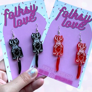 May include: A pair of black and red cat clock earrings with a white face and silver glitter. The earrings are on a pink card with the text "folksy love" and the website address "www.folksylove.com".