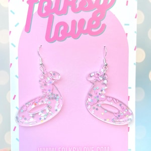 Può includere: Un paio di orecchini in resina rosa e trasparente con ganci argentati. Gli orecchini hanno la forma di una 'S' laterale con glitter incorporati nella resina. Gli orecchini sono esposti su una carta rosa con il testo 'folksy love' e 'www.folksylove.com' e 'TAG ME @folksylove'.