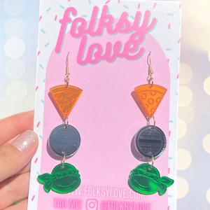 May include: A pair of dangle earrings featuring a pizza slice, a black and silver circle, and a green Teenage Mutant Ninja Turtle head. The earrings are on a white card with the text "folksy love" and "@folkslove".