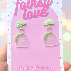 May include: A pair of pink and green glitter tequila bottle earrings with lime slices on top. The earrings are displayed on a pink and white card with the text "folksy love" and "www.folksylove.com".