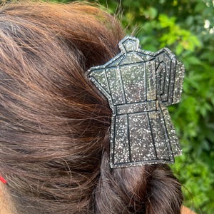 May include: A hair claw clip shaped like a moka pot coffee maker. The accessory is made of a translucent dark gray material embedded with numerous small silver glitter particles, featuring a distinct outline and a classic octagonal coffee pot silhouette.