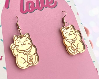 May include: Gold-colored acrylic earrings featuring a lucky cat design. The cats are holding a coin and have a smiling face. The earrings are hanging from gold-colored hooks.