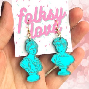 May include: A pair of turquoise blue earrings featuring a bust of a woman and a bust of a man. The earrings are on a white card with the text "folksy love" in pink and green letters.