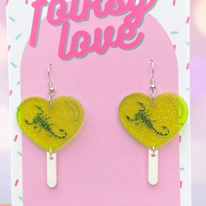 May include: A pair of green heart-shaped earrings with a scorpion inside. The earrings are on a pink background with the text "folksy love" and "www.folksylove.com" and "TAG ME! @folksylove".