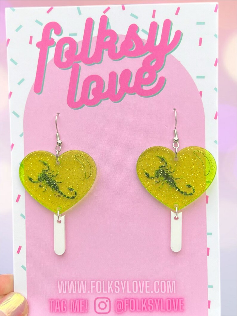 May include: A pair of green heart-shaped earrings with a scorpion inside. The earrings are on a pink background with the text "folksy love" and "www.folksylove.com" and "TAG ME! @folksylove".