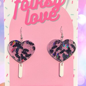 May include: A pair of pink heart-shaped earrings with a black bat pattern and iridescent glitter. The earrings are hanging from silver hooks and have a white dangle charm.