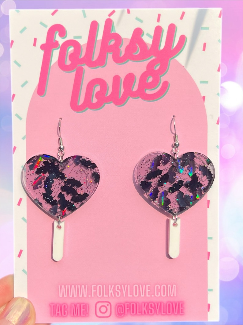 May include: A pair of pink heart-shaped earrings with a black bat pattern and iridescent glitter. The earrings are hanging from silver hooks and have a white dangle charm.