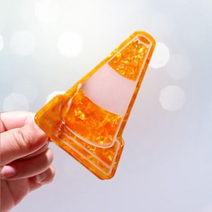 Peut inclure: Un objet décoratif en forme de cône de signalisation orange avec une texture pailletée. Le cône a une bande blanche et un intérieur rose. L'objet est tenu par une main sur un fond clair flou.