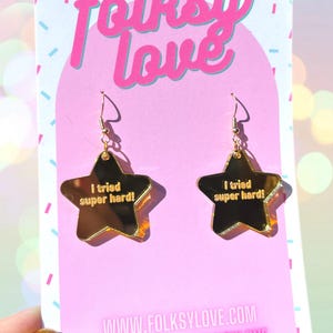 May include: Gold star-shaped earrings with the text "I tried super hard!" on each earring. The earrings are displayed on a pink background with the text "folksy love" and a website address.