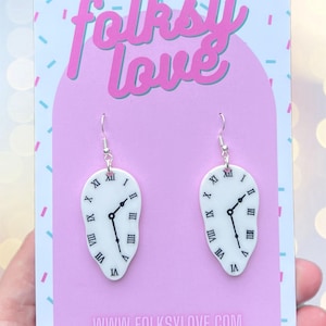 Puede incluir: Pendientes blancos con forma de reloj derritiéndose, con manecillas negras y números romanos. Cuelgan de ganchos plateados y se exhiben en una tarjeta rosa con el texto "folksy love" e información del sitio web.