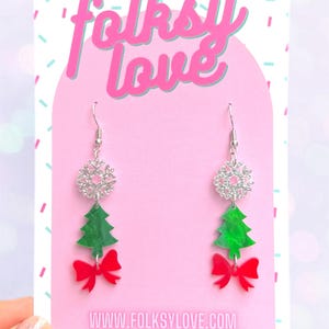 Puede incluir: Pendientes colgantes con copo de nieve plateado, árbol de Navidad verde y lazo rojo. Los pendientes están sobre un fondo rosa con el texto "folksy love" y "www.folksylove.com" y "TAG ME @folksylove".