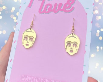 May include: Gold-coloured earrings featuring a robot face design. The earrings are on a pink card with the text "folksy love" and "www.folkslylove.com".