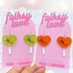 May include: A pair of heart-shaped earrings with a scorpion design. The earrings are made of clear resin with glitter and come in two colors: yellow and orange. The earrings are on a white card with the text "folksy love" and "www.folksylove.com".