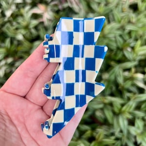 Puede incluir: Una pinza para el pelo con forma de rayo a cuadros azules y crema. El accesorio para el cabello presenta un vibrante patrón de cuadros azules y crema. La pinza se sostiene en una mano sobre un fondo de follaje verde.