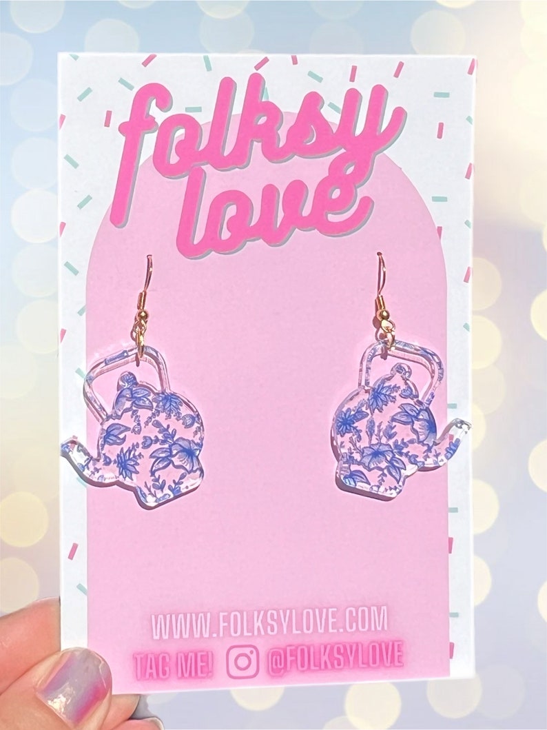 May include: A pair of clear acrylic earrings shaped like teapots with a blue and white floral pattern. The earrings are on a pink background with the text "folksy love" and "www.folkslove.com" and "TAG ME! @FOLKSLOVE".