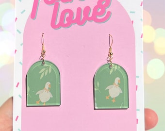 Puede incluir: Un par de pendientes de color verde y blanco con temática de ganso y ganchos dorados. Los pendientes tienen forma de ventanas arqueadas con un fondo verde y una ilustración de ganso blanco. El texto "folksy love" está impreso en la tarjeta.