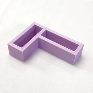 May include: A lilac-coloured, L-shaped organiser with two rectangular compartments. The item is made of a solid, smooth material and sits on a white surface. The design is simple and geometric, suitable for organising small items.