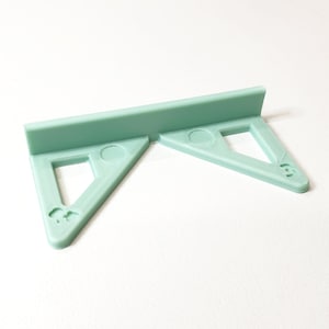 May include: A light green, 3D-printed corner shelf bracket. The bracket has two triangular supports with square cutouts and a rectangular back. Each triangle has a small, decorative design on the corner.