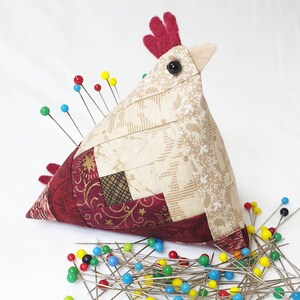 May include: A fabric pincushion shaped like a chicken, constructed from beige and dark red patterned quilt fabric pieces. It features a red felt comb, a beige beak, and a black bead eye, with numerous colourful sewing pins inserted.