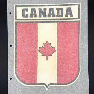 Op de afbeelding: Een vintage sticker met de Canadese vlag in rood en wit met een esdoornblad in het midden. De sticker is op een grijze achtergrond en heeft het woord "CANADA" in zwart bovenaan gedrukt.