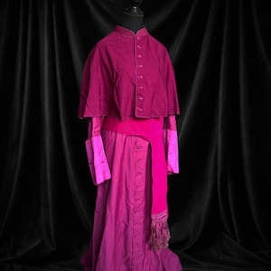 May include: A magenta-colored ecclesiastical garment set on a black mannequin. The set includes a button-down robe, a cape, and a sash. The cape and sash are a darker shade of magenta than the robe. The sash has a fringed end.