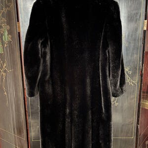 Puede incluir: Un abrigo largo de piel negro con cuello. El abrigo est&aacute; colgado en una percha y est&aacute; delante de una pantalla negra y dorada.