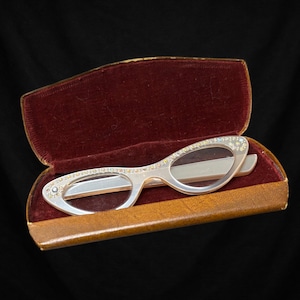 Puede incluir: Gafas plateadas de ojo de gato con lentes transparentes, adornadas con pedrería en el borde superior. Las gafas están en un estuche vintage abierto, con exterior marrón e interior de terciopelo rojo, sobre un fondo negro.