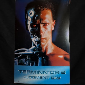 May include: A VHS tape for the film "Terminator 2: Judgment Day." The cover shows a cyborg with a metallic endoskeleton on one side of its face and a human face on the other. The title is in a blue, metallic font.
