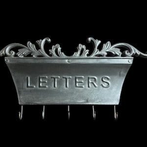 May include: A silver-coloured metal wall-mounted letter holder with the word "LETTERS" embossed on the front. It features decorative scrollwork at the top and four hooks below for keys. The background is black.