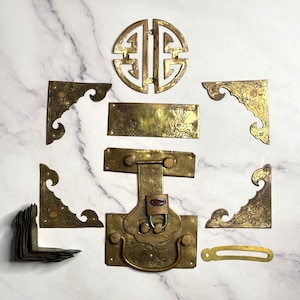 May include: An assortment of antique brass hardware components, including corner brackets, a circular decorative piece, a rectangular plate, and a trunk latch with a red lock. The pieces have ornate floral and scrollwork designs.
