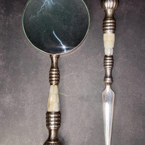 May include: A silver magnifying glass and letter opener set. The magnifying glass has a round lens and a decorative handle with a mother-of-pearl inlay. The letter opener has a similar handle and a pointed blade. The set is displayed on a dark background.