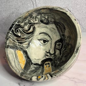 May include: A ceramic bowl with a hand-painted portrait of a person. The portrait is in black, white, and yellow. The bowl has a gray and white marbled effect around the rim and exterior. The bowl is a unique piece of art.