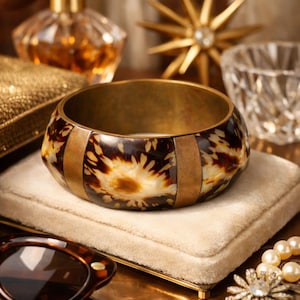 Puede incluir: Un brazalete ancho y decorativo con un patrón de carey y detalles metálicos dorados. El brazalete se encuentra sobre una superficie de terciopelo color crema, rodeado de otros accesorios vintage.